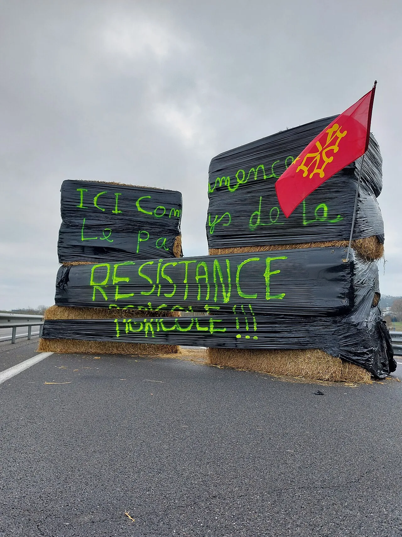 Barricade de grosses bottes de paille emballées de plastique noir sur une route, peintes en vert « Ici commence le pays de la résistance agricole », surmontée d'un drapeau occitan.