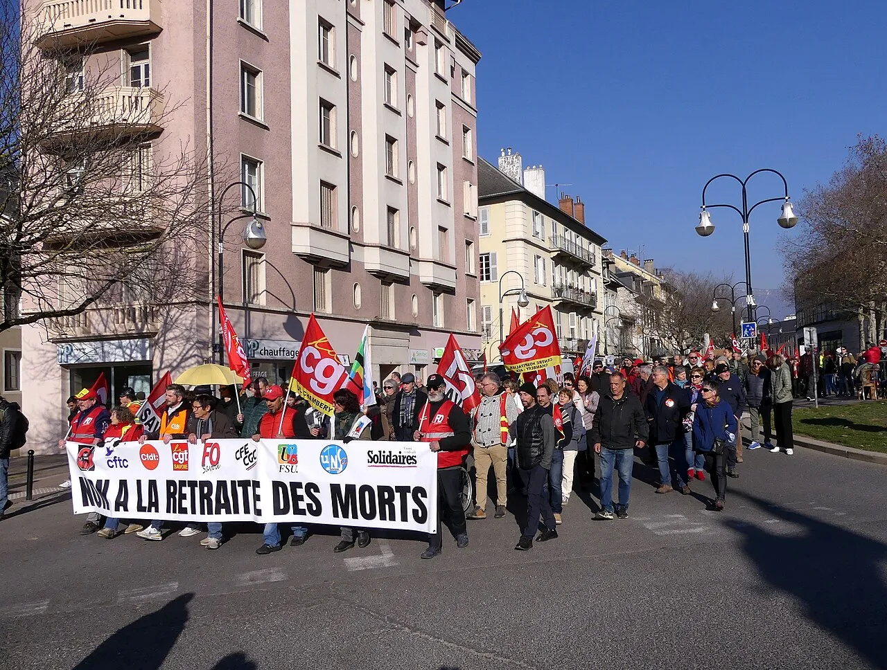 Tête de cortège dans une rue ensoleillée : une banderole blanche porte les logos de plusieurs syndicats et le slogan « Non à la retraite des morts », suivie de drapeaux CGT et FO.