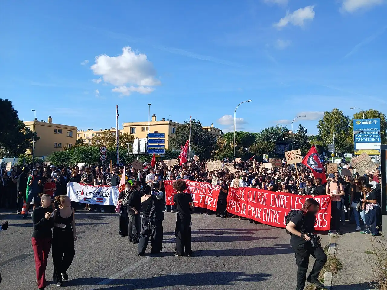 Cortège de manifestants, en majorité jeunes, sur une large avenue ensoleillée, derrière des banderoles rouges et des pancartes en carton.