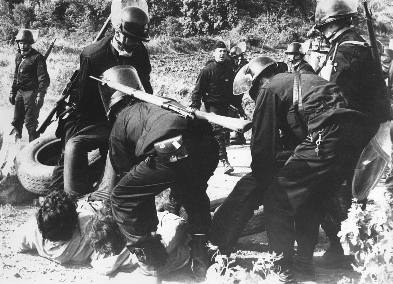 Photographie en noir et blanc de gendarmes casqués, fusil à l'épaule, maintenant des manifestants au sol près de pneus.