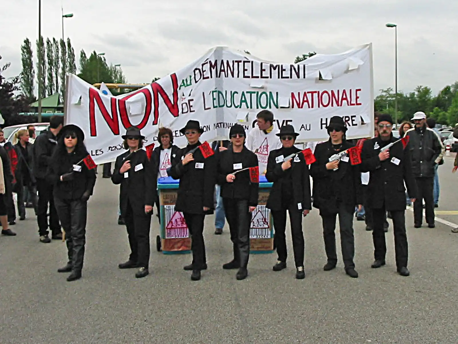 Cortège derrière une grande banderole blanche « Non au démantèlement de l'Éducation nationale » ; au premier rang, des manifestants vêtus de noir, chapeau et lunettes, tiennent des pistolets en plastique et de petits drapeaux rouges.