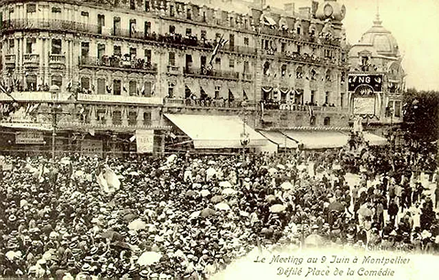 Carte postale sépia : une foule très dense, coiffée de chapeaux, remplit une place bordée d'immeubles à balcons pavoisés ; des ombrelles blanches dépassent çà et là.