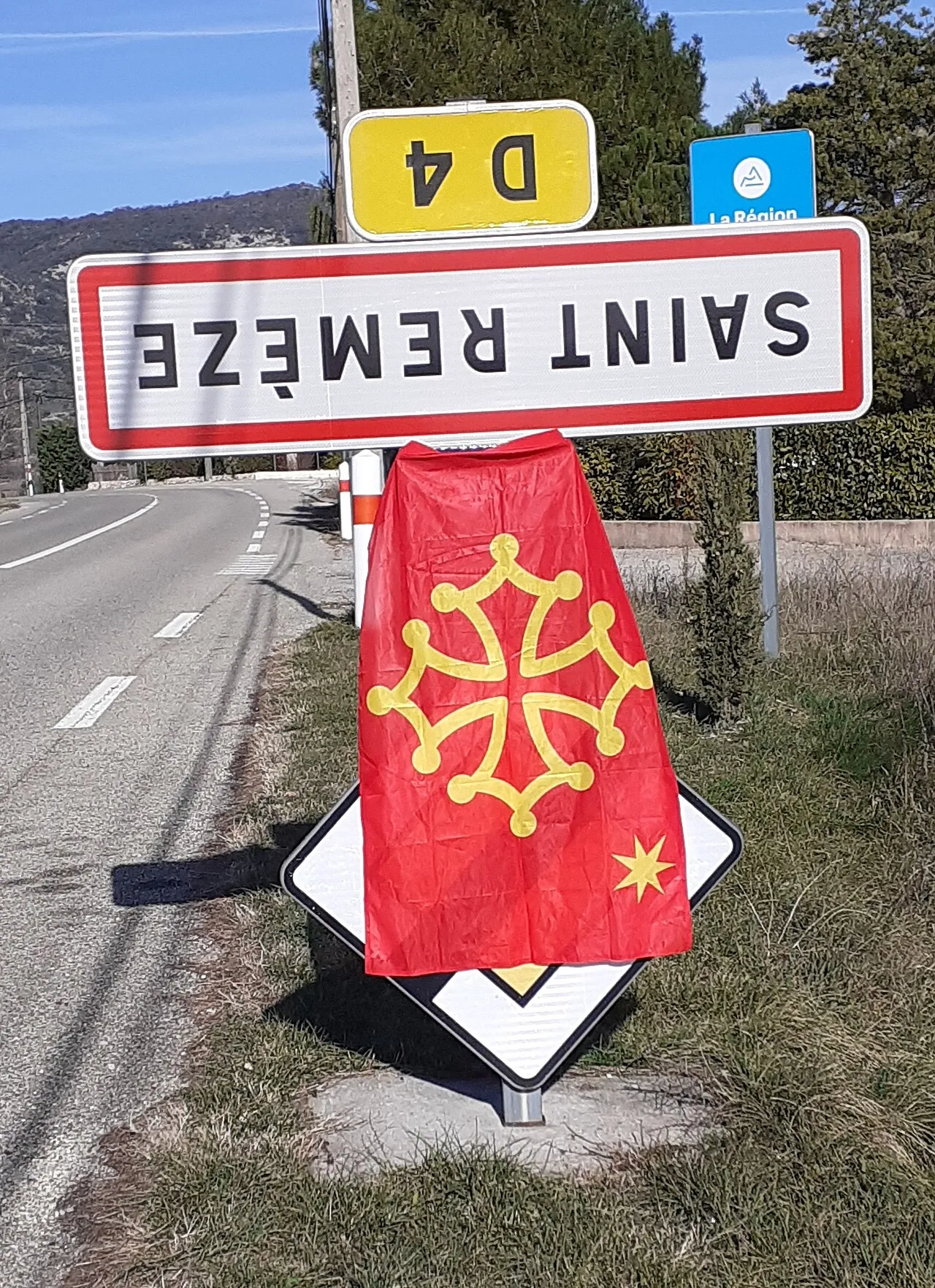 Panneau d'entrée de la commune de Saint-Remèze fixé à l'envers, avec un drapeau occitan accroché dessous, au bord d'une route départementale.