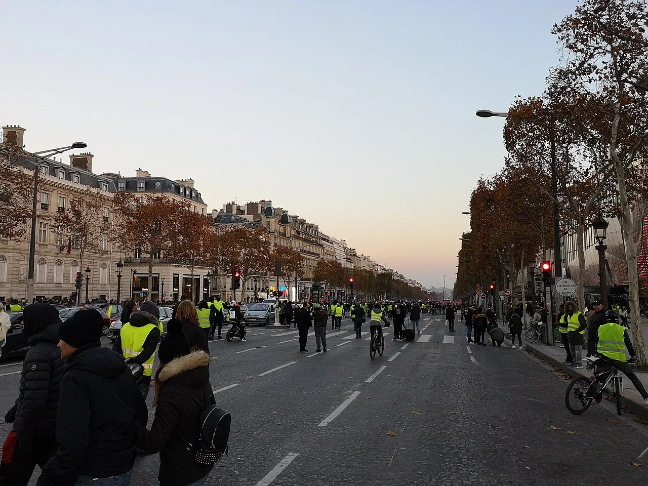 Avenue des Champs-Élysées au crépuscule, presque sans circulation, parcourue par des piétons dont beaucoup portent un gilet jaune ; quelques voitures à gauche.