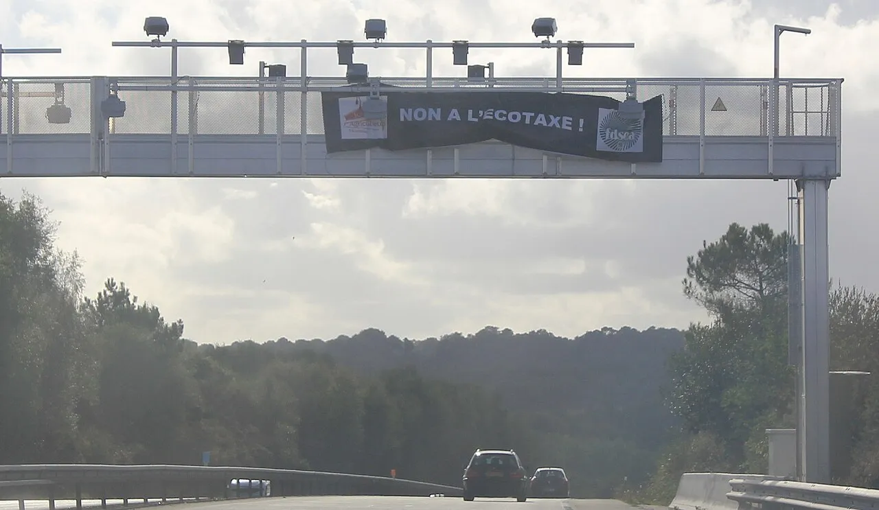 Portique métallique au-dessus d'une voie rapide, équipé de caméras, portant une banderole noire « Non à l'écotaxe ! » ; deux voitures passent dessous.